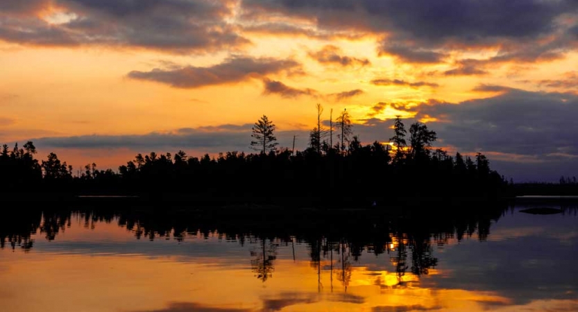 the sunset is reflected on a calm lake alongside a tree-lined shore